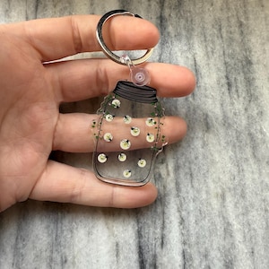 Puede incluir: Un llavero de acrílico transparente con forma de tarro de cristal lleno de luciérnagas brillantes. El tarro tiene una tapa negra y está rodeado de enredaderas verdes.