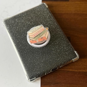 Könnte beinhalten: Eine schwarze Tablet-Hülle mit transparentem, glitzerndem Finish. Ein klarer, runder Telefonhalter mit einem Stapel illustrierter Bücher ist angebracht. Die Bücher sind in Orange-, Grün- und Beigetönen gehalten, mit dem Text "Harvest Season".