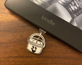 May include: A black Kindle e-reader with the word "kindle" printed on the front. A white bucket with a smiling face is attached to the bottom of the e-reader.