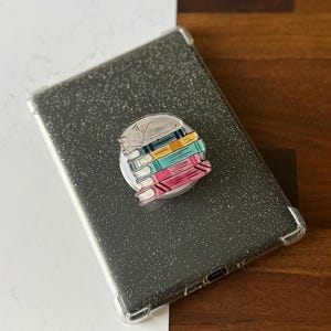 May include: A glittery gray tablet case with a clear bumper. A round phone grip is attached, featuring a sleeping gray cat on a stack of colorful books. The books are pink, teal, yellow, and blue.