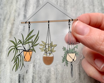 May include: A clear sticker with a design of three hanging plants in macrame planters. The plants are green and brown, and the planters are beige, brown, and white.
