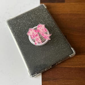 Puede incluir: Una funda transparente para tableta con una parte trasera gris oscuro brillante. Un agarre para teléfono en forma de lazo rosa con un estampado floral está adherido a la tableta. El lazo tiene un fondo blanco y un diseño floral rosa.