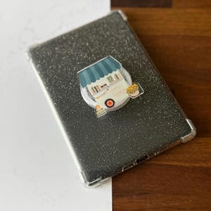 May include: A black glitter tablet case with a clear bumper. A pop-up phone grip is attached, featuring a whimsical illustration of a coffee and books cart with the text "Coffee Books". The cart has a blue awning and a red wheel.