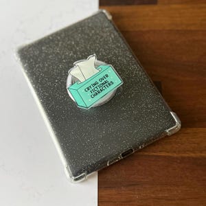 May include: A clear phone case with a glittery, speckled design. A pop-up phone grip is attached, featuring a teal box of tissues with the text "Crying Over Fictional Characters."