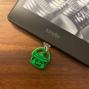 May include: A green plastic bucket charm with a smiling jack-o'-lantern face attached to a black Kindle e-reader.