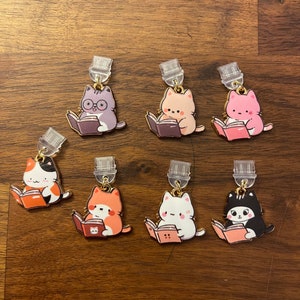 May include: Set of seven adorable cat bookmarks. Each bookmark features a different colored cat reading a book. The cats are all wearing different expressions and have different colored fur. The bookmarks are made of metal and have a gold finish.