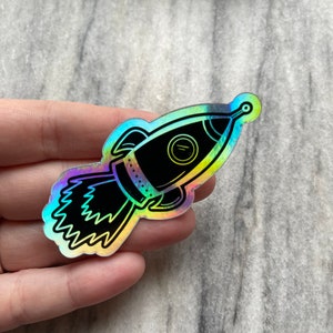 Op de afbeelding: Een holografische sticker van een zwart-witte raket met een regenboogkleurige, iriserende achtergrond.