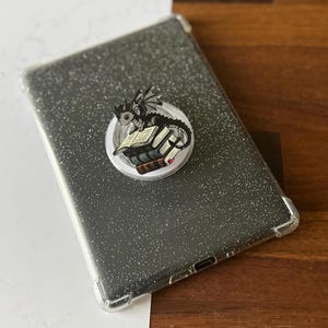 May include: A clear, glittery tablet case with a dragon-themed pop-up grip. The pop-up grip features a black and gray dragon perched atop a stack of books. The tablet case is gray and has a clear border.