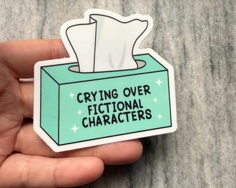 May include: A sticker featuring a teal tissue box with white tissues peeking out. The text "CRYING OVER FICTIONAL CHARACTERS" is printed in black. White stars are on the teal box. The sticker is held in a hand.