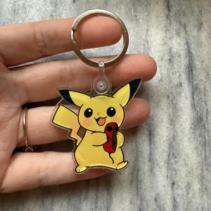May include: A yellow Pikachu keychain holding a red and black video game controller. The keychain is made of clear plastic and has a silver metal ring.