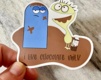 May include: A sticker featuring two cartoon characters, one blue and one yellow, with chocolate-colored accents. The blue character has a sad expression, while the yellow character has a wide smile. The sticker includes the text "i like chocolate milk".