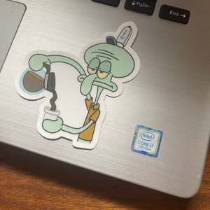 May include: A sticker featuring Squidward from Spongebob Squarepants pouring coffee into a cup. The sticker is on a laptop. Squidward is light green with a brown shirt and a sailor hat. The laptop has a silver finish and a Core i7 sticker.