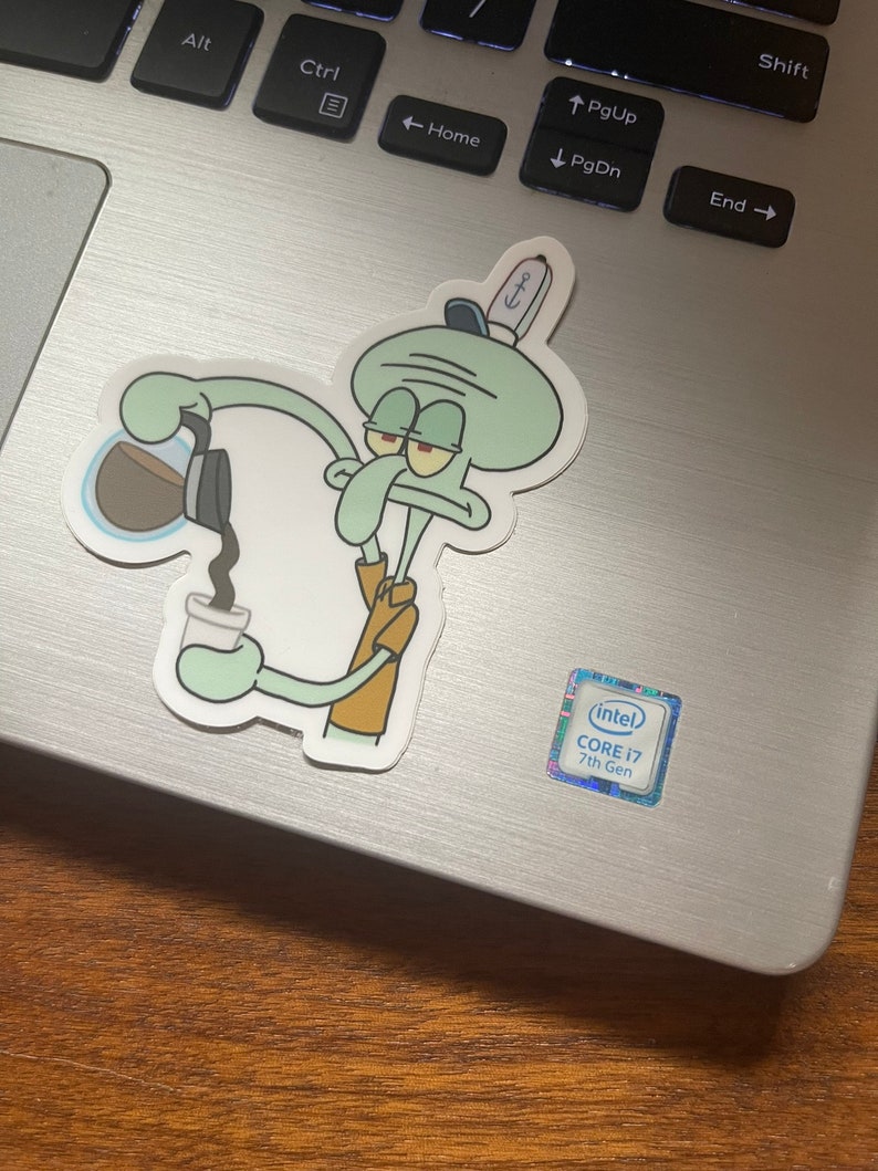 May include: A sticker featuring Squidward from Spongebob Squarepants pouring coffee into a cup. The sticker is on a laptop. Squidward is light green with a brown shirt and a sailor hat. The laptop has a silver finish and a Core i7 sticker.