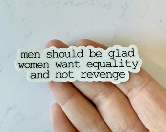 May include: A white sticker with black text that reads "men should be glad women want equality and not revenge." The sticker is rectangular with rounded corners. The sticker is held in a hand.