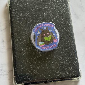 May include: A black tablet case with a silver glitter finish and reinforced clear corners. A circular phone grip is attached to the center, featuring a moth illustration reading a green book with the text LIBRARY LURKER BOOK CLUB in pink.