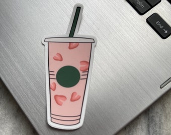 May include: A pink and white sticker of a coffee cup with a green logo and a straw. The cup is filled with pink hearts.