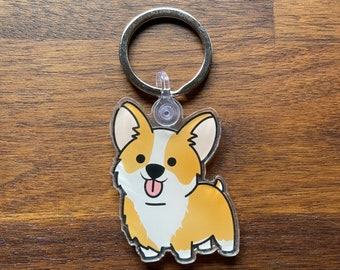 Op de afbeelding: Een heldere acryl sleutelhanger met een cartoon corgi hond met een vrolijke uitdrukking. De corgi heeft een witte en oranje vacht, met een roze tong die uitsteekt. De sleutelhanger heeft een zilveren metalen ring.