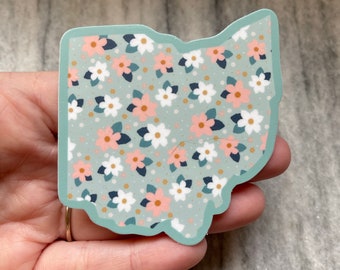 May include: A sticker of the state of Ohio with a floral pattern. The sticker is light blue with white and pink flowers and gold accents.