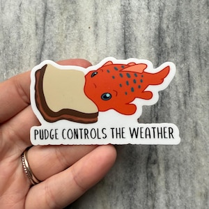 May include: A sticker featuring a cartoon orange fish with blue spots biting into a piece of bread. The text "PUDGE CONTROLS THE WEATHER" is below the image. The sticker is held by a hand.