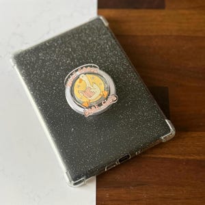 May include: A black glitter tablet case with a clear bumper. A circular pin with the text "Silly Goose Book Club" and a cartoon goose reading a book is attached to the case.
