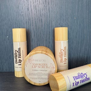 Puede incluir: Tres tubos de madera con etiquetas que dicen "Buttery Lip Balm" en una variedad de colores. Los tubos est&aacute;n al lado de un frasco de madera con una etiqueta que dice "Smokers Lip Scrub" y una descripci&oacute;n del producto.