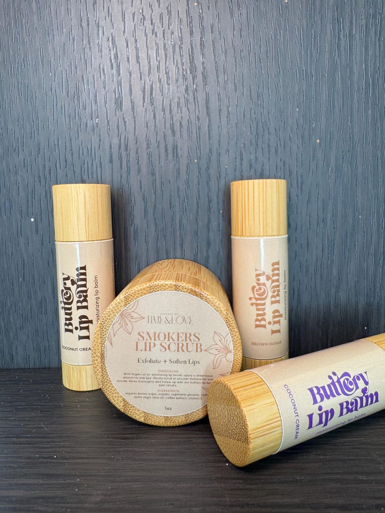 Puede incluir: Tres tubos de madera con etiquetas que dicen "Buttery Lip Balm" en una variedad de colores. Los tubos est&aacute;n al lado de un frasco de madera con una etiqueta que dice "Smokers Lip Scrub" y una descripci&oacute;n del producto.
