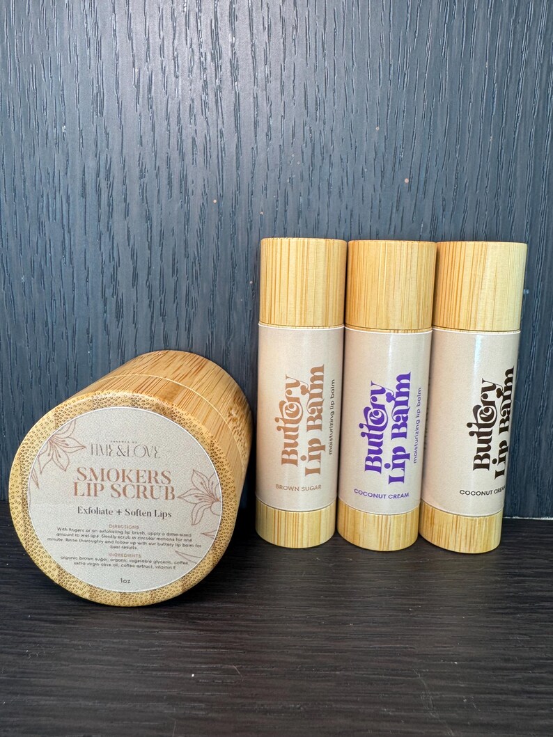 May include: Three brown sugar, coconut cream, and cocoa lip balms in bamboo tubes with the text "Buttery Lip Balm" and "Moisturizing Lip Balm". A brown bamboo jar with the text "Smokers Lip Scrub" and "Exfoliate + Soften Lips".