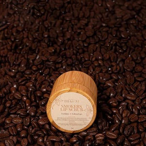 Puede incluir: Un peque&ntilde;o tarro de madera de exfoliante labial con una etiqueta blanca que dice "Time & Love Smokers Lip Scrub Exfoliate + Soften Lips". El tarro est&aacute; sentado sobre un lecho de granos de caf&eacute;.