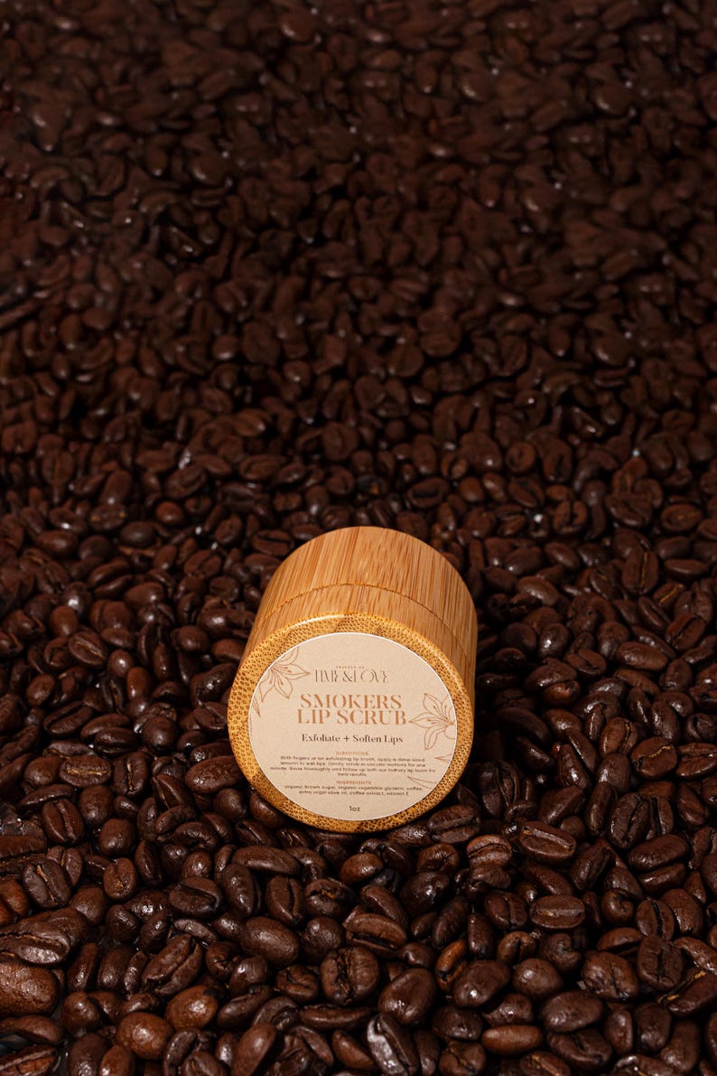 Puede incluir: Un peque&ntilde;o tarro de madera de exfoliante labial con una etiqueta blanca que dice "Time & Love Smokers Lip Scrub Exfoliate + Soften Lips". El tarro est&aacute; sentado sobre un lecho de granos de caf&eacute;.
