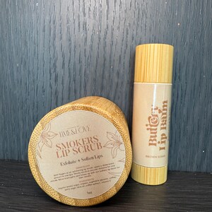 Puede incluir: Un conjunto de dos recipientes de madera para productos de cuidado de labios. El recipiente m&aacute;s grande es un exfoliante labial etiquetado como "Smokers Lip Scrub" con el texto "Exfoliate + Soften Lips". El recipiente m&aacute;s peque&ntilde;o es un b&aacute;lsamo labial etiquetado como "Butio Lip Balm" con el texto "Brown Sugar".