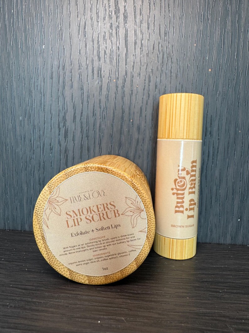 Puede incluir: Un conjunto de dos recipientes de madera para productos de cuidado de labios. El recipiente m&aacute;s grande es un exfoliante labial etiquetado como "Smokers Lip Scrub" con el texto "Exfoliate + Soften Lips". El recipiente m&aacute;s peque&ntilde;o es un b&aacute;lsamo labial etiquetado como "Butio Lip Balm" con el texto "Brown Sugar".