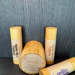 May include: Three wooden tubes with labels that read "Buttery Lip Balm" in a purple and white font. The tubes are in a variety of colours and have different scents, including coconut cream and brown sugar. The tubes are sitting on a dark wooden surface. There is also a wooden jar with a label that reads "Smokers Lip Scrub" in a brown font. The jar is sitting on a dark wooden surface.