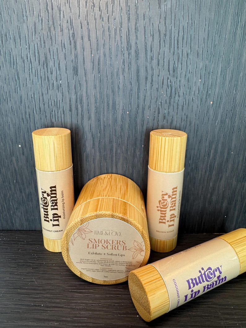 May include: Three wooden tubes with labels that read "Buttery Lip Balm" in a purple and white font. The tubes are in a variety of colours and have different scents, including coconut cream and brown sugar. The tubes are sitting on a dark wooden surface. There is also a wooden jar with a label that reads "Smokers Lip Scrub" in a brown font. The jar is sitting on a dark wooden surface.