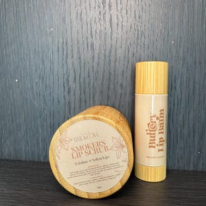 Puede incluir: Dos recipientes de madera con etiquetas. El recipiente m&aacute;s grande es un exfoliante labial etiquetado como "Smokers Lip Scrub" y el recipiente m&aacute;s peque&ntilde;o es un b&aacute;lsamo labial etiquetado como "Buttery Lip Balm". Ambos recipientes est&aacute;n hechos de madera natural y tienen un color marr&oacute;n claro.
