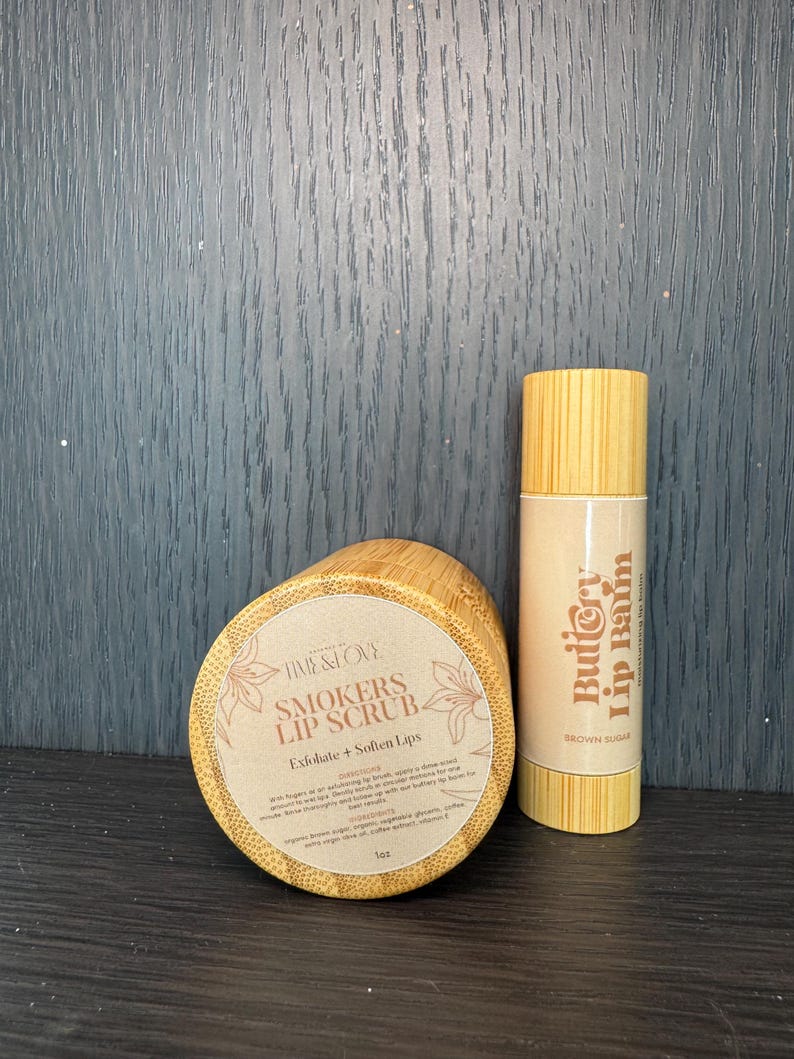 Puede incluir: Dos recipientes de madera con etiquetas. El recipiente m&aacute;s grande es un exfoliante labial etiquetado como "Smokers Lip Scrub" y el recipiente m&aacute;s peque&ntilde;o es un b&aacute;lsamo labial etiquetado como "Buttery Lip Balm". Ambos recipientes est&aacute;n hechos de madera natural y tienen un color marr&oacute;n claro.