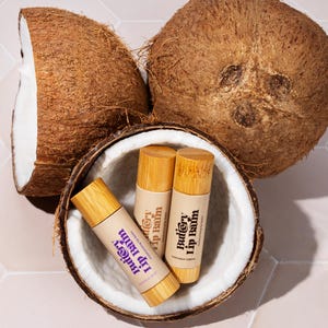 May include: Three wooden lip balm tubes with the text "Buttery Lip Balm" on the label. The tubes are inside of a halved coconut shell.