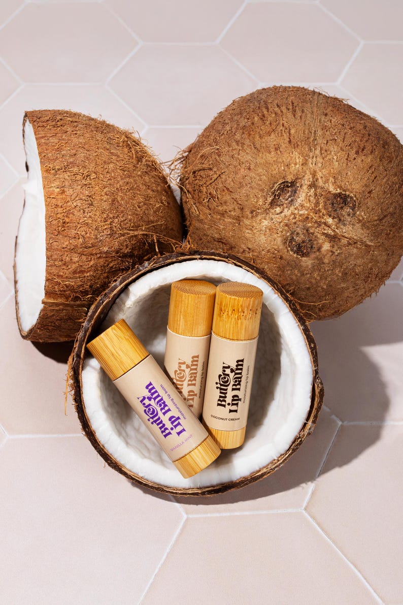May include: Three wooden lip balm tubes with the text "Buttery Lip Balm" on the label. The tubes are inside of a halved coconut shell.