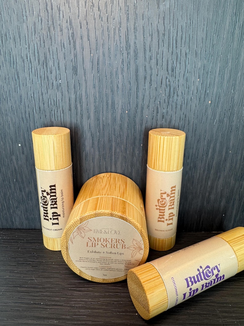 Puede incluir: Tres tubos de madera con etiquetas que dicen "Buttery Lip Balm" en letras moradas y blancas. Los tubos son de bamb&uacute; y tienen un acabado de madera natural. El tubo del medio es un frasco con una etiqueta que dice "Time & Love Smokers Lip Scrub Exfoliate + Soften Lips".