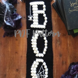Puede incluir: Una pared colgante de crochet en blanco y negro con la palabra "BOO" escrita en pompones de hilo blanco. La "O" tiene un diseño de telaraña blanca. La pared colgante tiene borlas blancas en la parte inferior. El texto "PDF Pattern" es visible en la imagen.