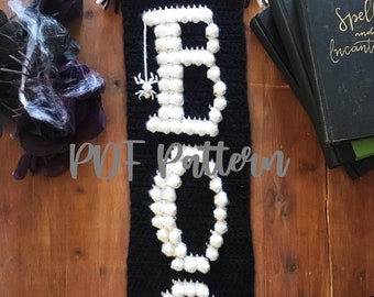 Può includere: Un'appendice da parete in crochet bianco e nero con la parola "BOO" scritta in pompon di lana bianca. La "O" ha un disegno di ragnatela bianca. L'appendice ha frange bianche in basso. Il testo "PDF Pattern" è visibile nell'immagine.