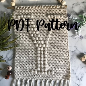 Puede incluir: Un tapiz de pared de crochet beige con una gran 'Y' de hilo blanco texturizado en el centro. El tapiz tiene un borde de flecos y está sujeto a una varilla de madera con una cuerda.