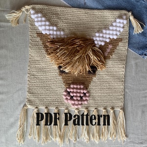 Pode incluir: Padrão de crochê para uma tapeçaria de parede de vaca das terras altas. O padrão é bege com detalhes marrons e rosados. A peça acabada tem franjas brancas e bege.