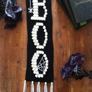 May include: A black and white crocheted wall hanging with the word "BOO" spelled out in white yarn. The "O" has a spider web design and a small spider. The hanging has white tassels at the bottom.