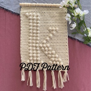 May include: Beige crochet wall hanging with a letter "K" design, featuring a wooden dowel at the top and fringe at the bottom. The text "PDF Pattern" is visible at the bottom. This handmade piece adds a touch of bohemian style.