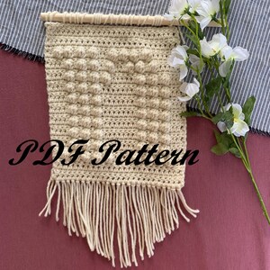 May include: A beige crochet wall hanging with a textured pattern and fringe. The hanging is made with a chunky yarn and has a simple, modern design. The text "PDF Pattern" is written in black on the hanging.