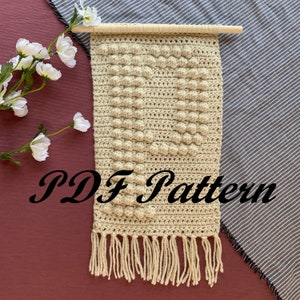 May include: A beige crochet wall hanging with a heart shape and a fringe border. The hanging is attached to a wooden dowel rod. The text "PDF Pattern" is visible in the image.