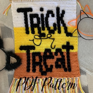 May include: Crocheted wall hanging with the words "Trick or Treat" in black yarn. The hanging is made with white, yellow, and orange yarn and has a fringe at the bottom. The text "PDF Pattern" is written in black on the bottom of the hanging.