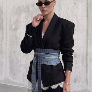 Pode incluir: Um blazer preto com uma faixa larga, azul e cinza estampada amarrada na cintura. O blazer tem decote em V e é usado sobre um top de renda branca. A pessoa está usando óculos de sol pretos e pulseiras de prata.
