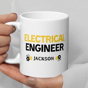 May include: White ceramic mug featuring the text "ELECTRICAL ENGINEER" in yellow and black, with the name "JACKSON" below. The mug has a handle and gear icons on either side of the name.