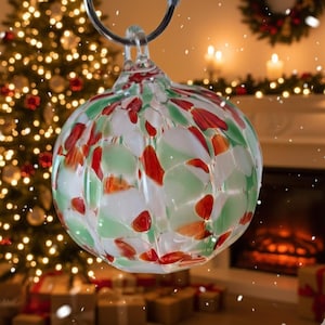 Puede incluir: Una bola de Navidad redonda de vidrio con un diseño moteado en rojo, blanco y verde. La bola está suspendida por un lazo de metal plateado. El fondo presenta un árbol de Navidad decorado y una chimenea.